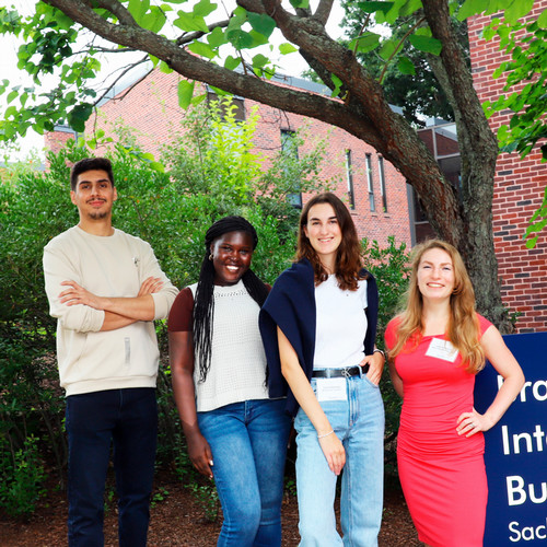 The four Peace Scholars pose outside the Lemberg building in front of some trees and next to a blue Brandeis International Business School sign.