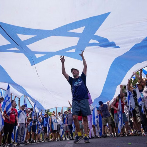 A man stands with his arms raised under a large Israeli flag.