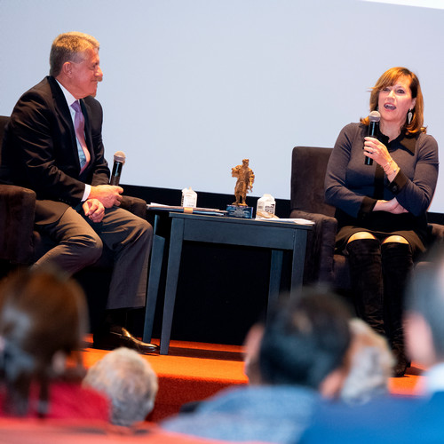 Jean Hynes speaks into a microphone while seated onstage next to Perry Traquina with a bronze statue of Louis Brandeis on a table between them.