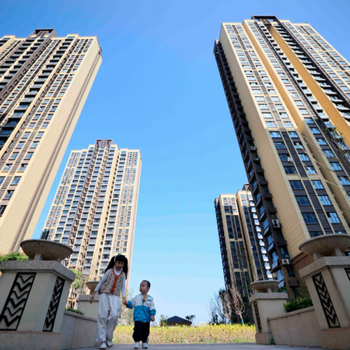 A shot looking up at four tall apartment towers with a woman and small child walking in front of the entrance to the courtyard of the complex.