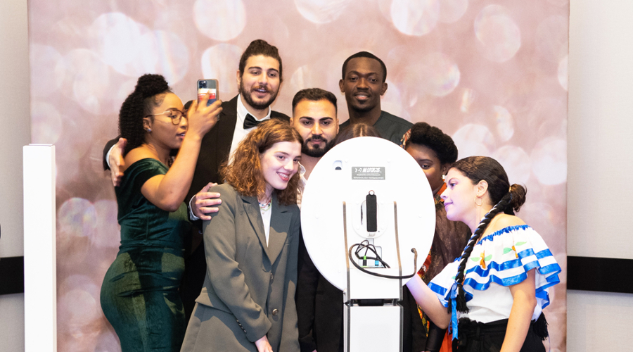 A group of students pose in front of a camera with a pink shimmering background behind them.