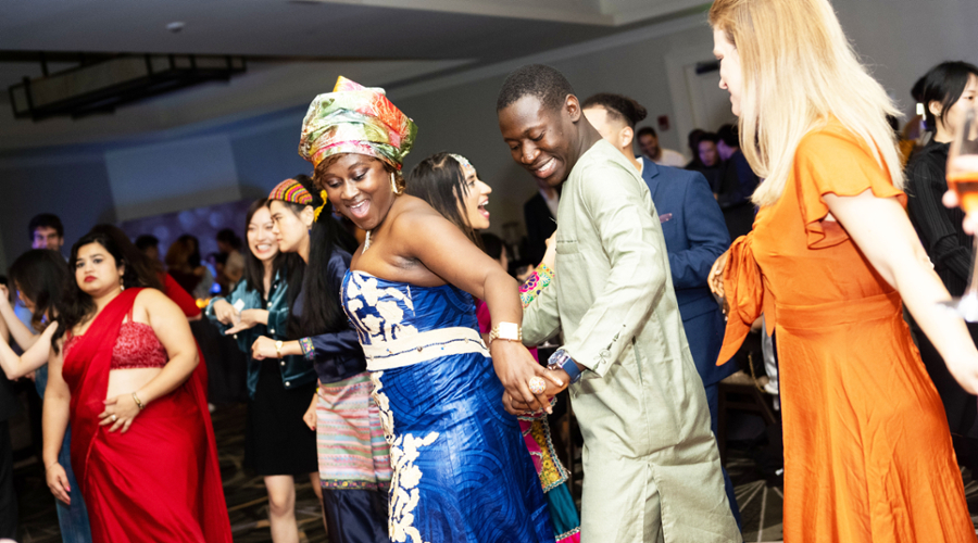 A student in a blue and white dress with a colorful headdress dances in front of another student wearing a light green shirt.