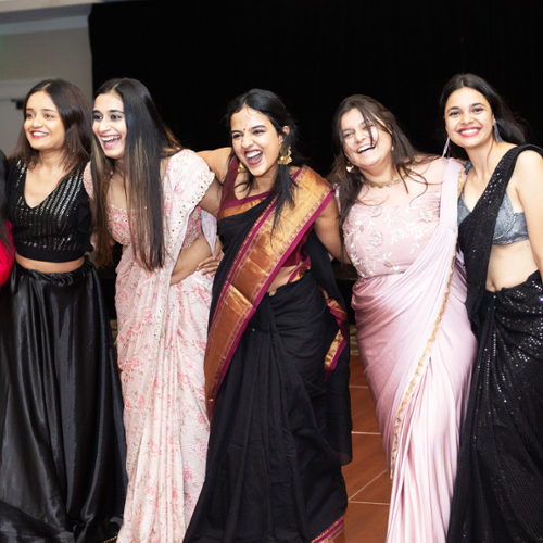 Bhavya Vishishta Kondamuri in a black saree with a gold sash laugh with fellow students in Indian dress at the Global Gala.