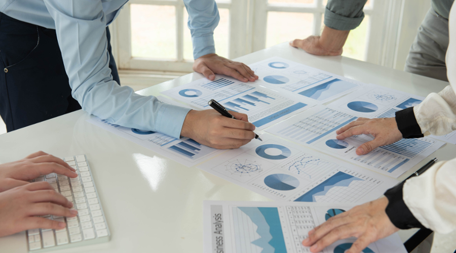 Papers with charts on them lie scattered on a table with people leaning over in discussion.