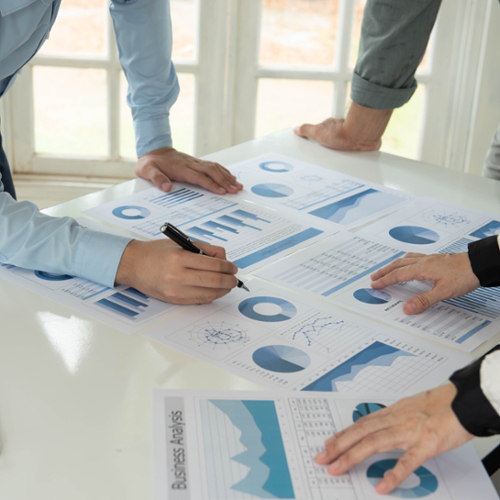 Papers with charts on them lie scattered on a table with people leaning over in discussion.