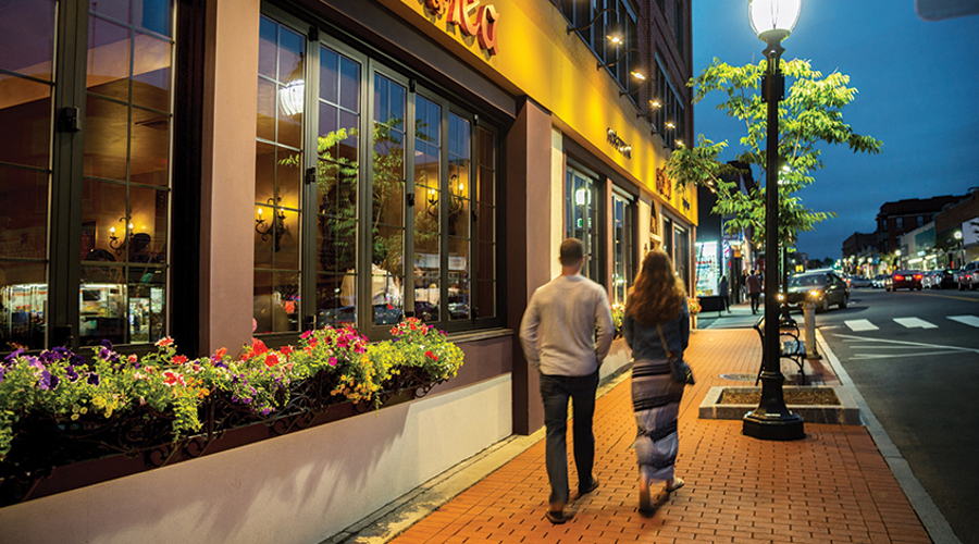 A shot of a couple walking together down Moody Street past a lamppost at dusk.