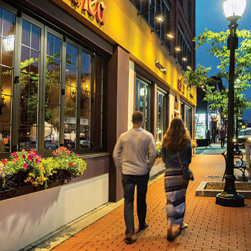 A shot of a couple walking together down Moody Street past a lamppost at dusk.