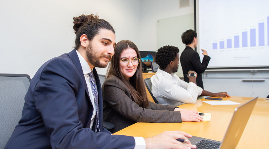 Students in professional dress working together on a laptop while another student examines a bar graph on a presentation slide.