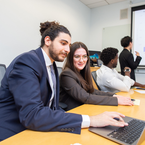 Students in professional dress working together on a laptop while another student examines a bar graph on a presentation slide.