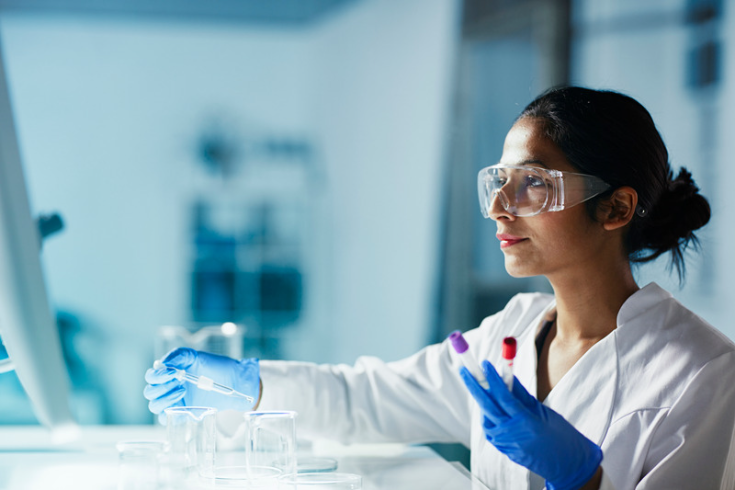 Woman in white lab coat and goggles holds test tubes while looking at computer