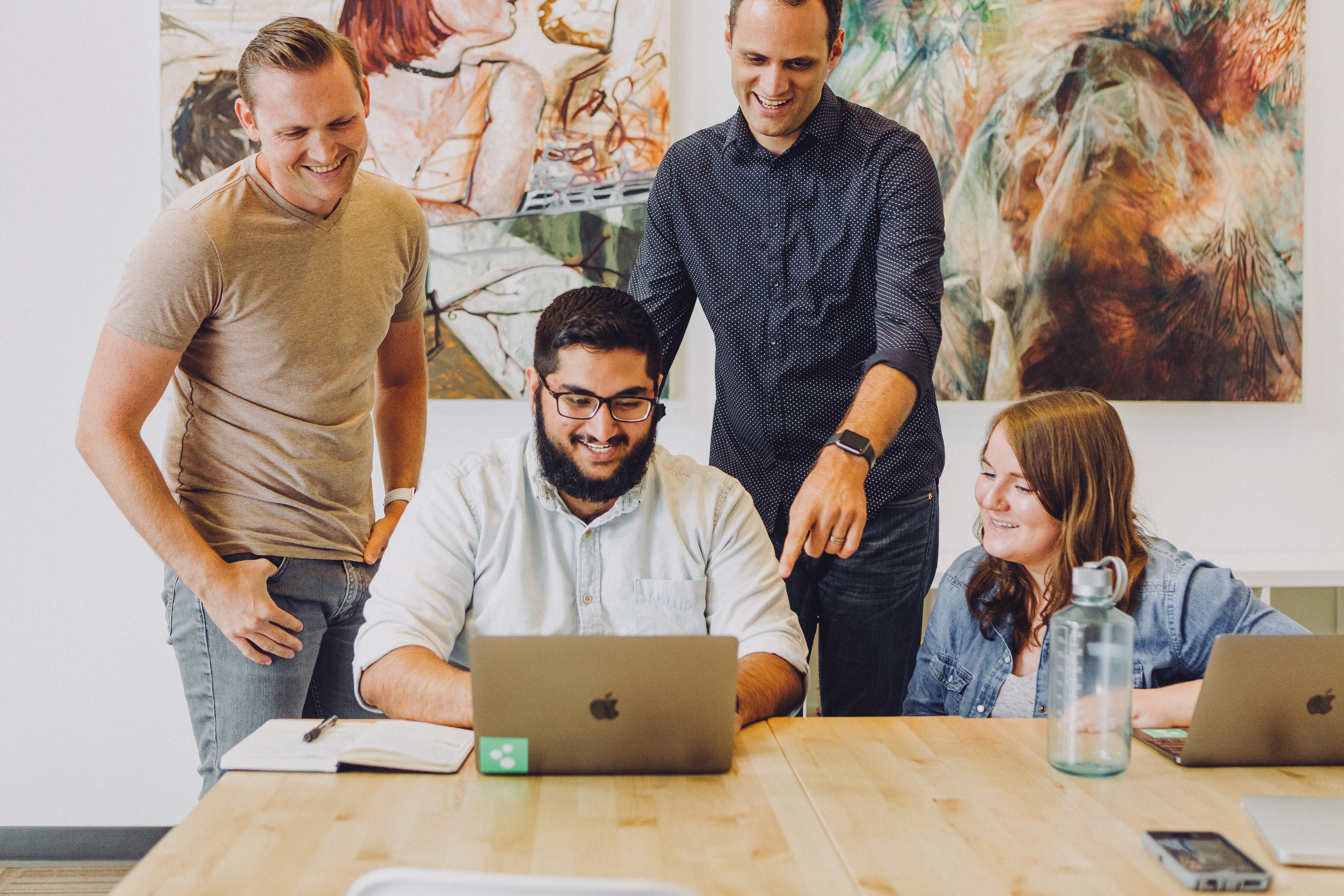 Four people, two standing and two sitting, look at a laptop screen together while smiling