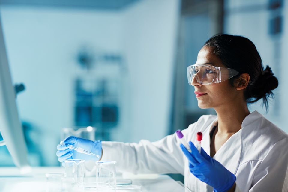 Woman in lab coat holds test tubes of colored liquid while looking at computer screen