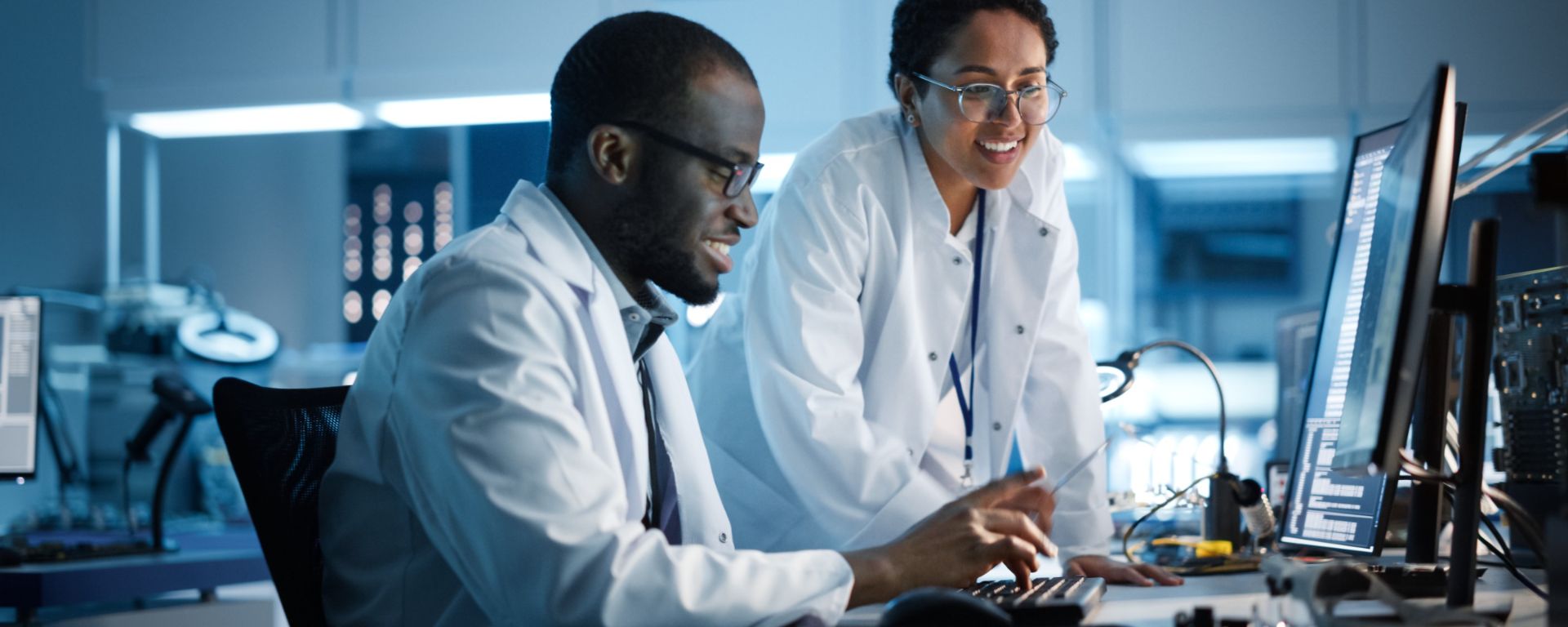 Two people in a science lab look at a computer, smiling, while one types on a keyboard