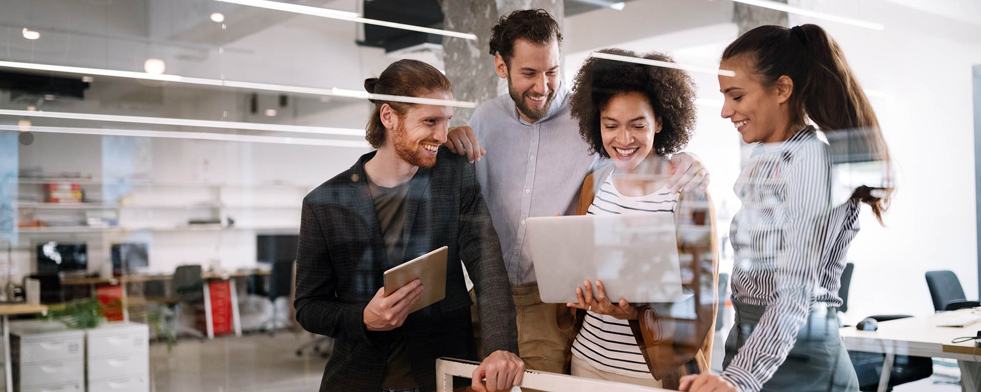 Two women and two men are clustered together as one of the women shows them something on her laptop with them. All are smiling.