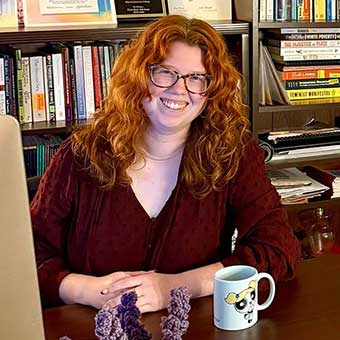 Anne Marie Foley sits at a desk in front of shelves with lots of books and walls with colorful art.