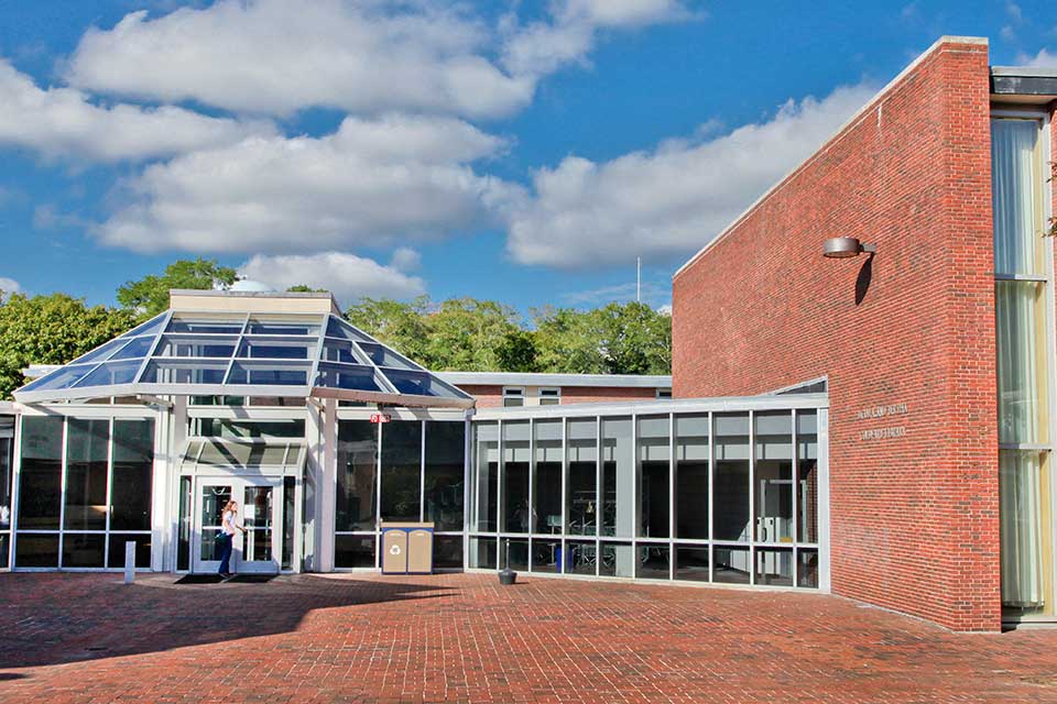 A sunny day with a student entering the glass door entrance to the Goldfarb Library.  The library is a brick building with brick patio entryway. 