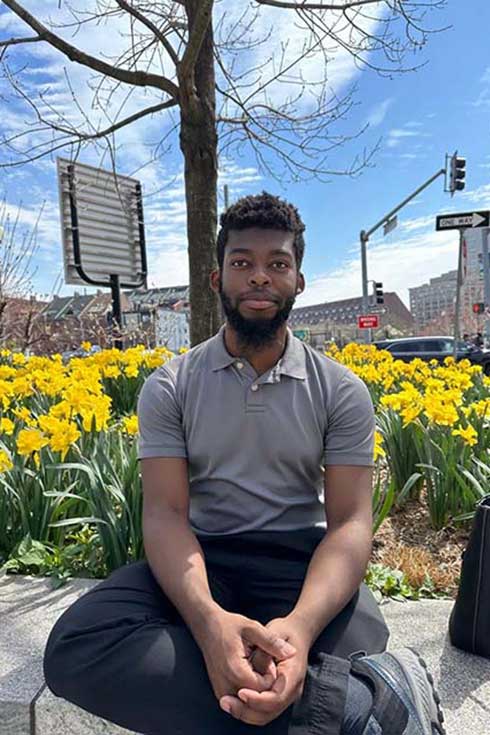 Abichael Belizaire sits on a stone wall in front of flowers in a city setting.