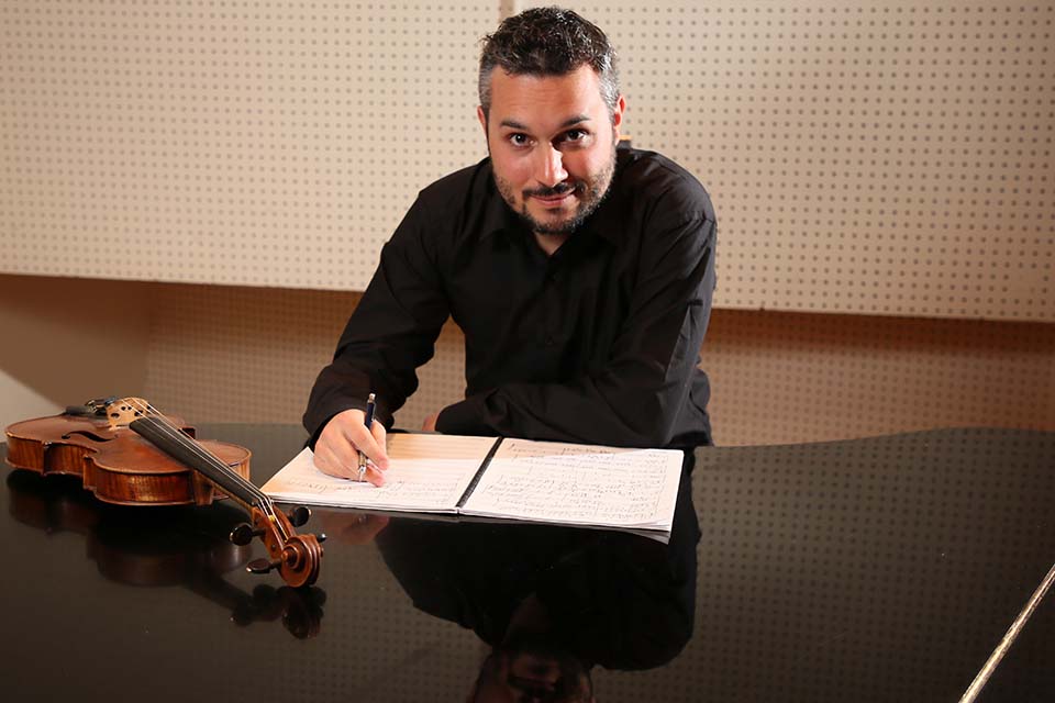 Ali Puskulcu leans on a piano, writing on a music score. There is a violin on the piano lid, and Ali is reflected in the lid's surface.