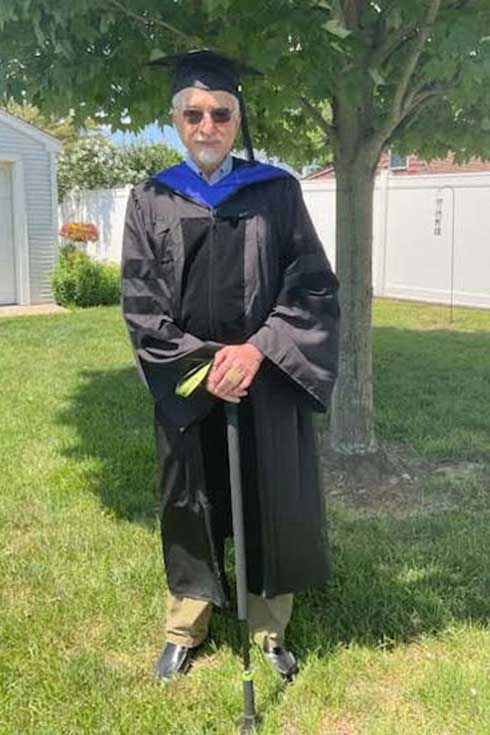 Bill Abbate, wearing graduation robes and holding a cane, stands in front of a tree.