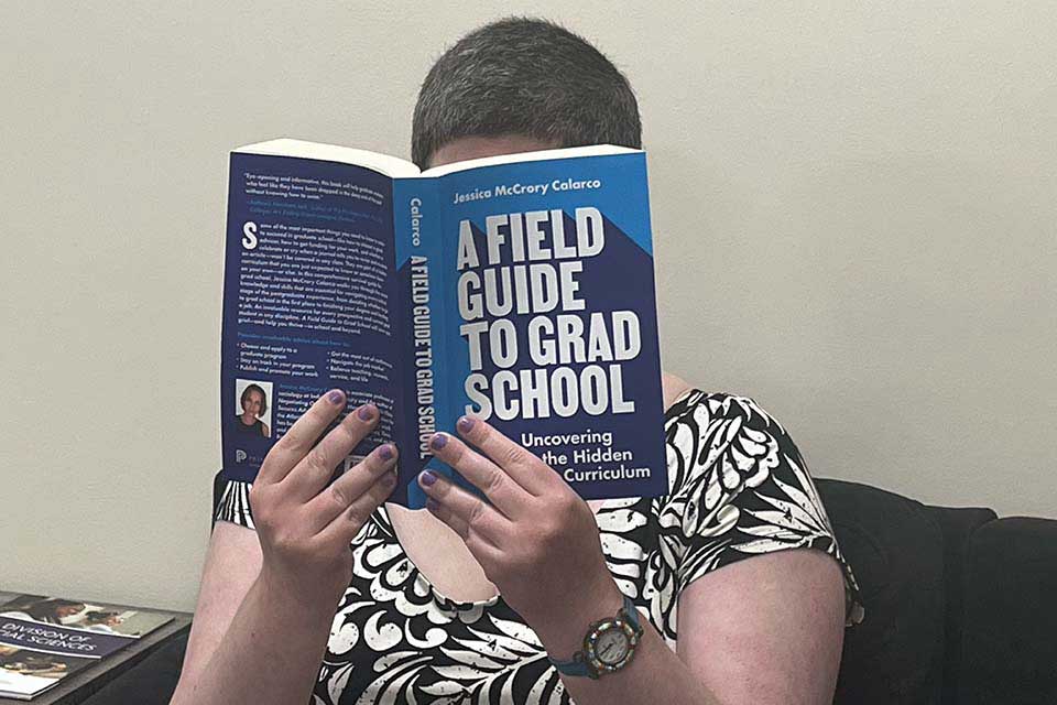A woman sits in a chair, holding an open copy of A Field Guide to Grad School in front of her face.