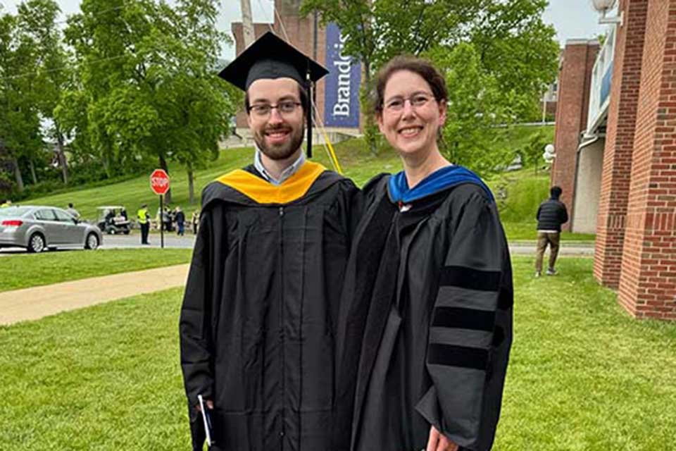 Josh Broderick Phillips and Lotus Goldberg stand on the Brandeis campus wearing academic regalia.