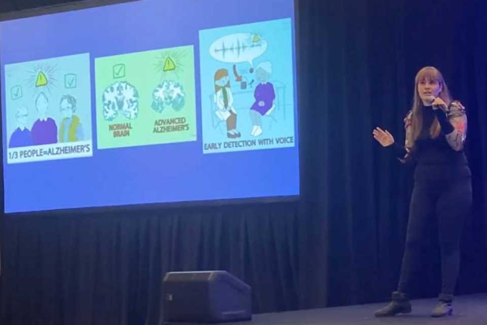 Liz Mahon speaks into a microphone at 3MT Nationals, next to her slide.