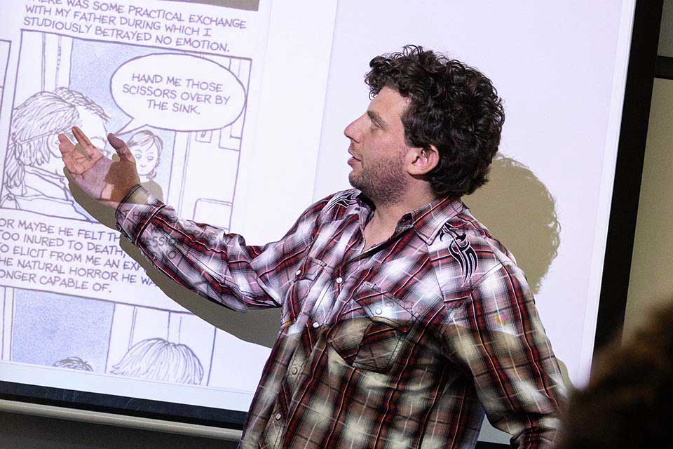 Rafi Abrahams stands in front of a screen, pointing at a panel from Alison Bechdel's comic Fun Home.