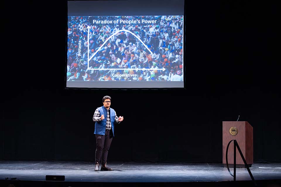Arjun Kumar Singh presents onstage in front of a large slide reading "Paradox of People's Power" and showing a curving graph in front of a photo of a large crowd.