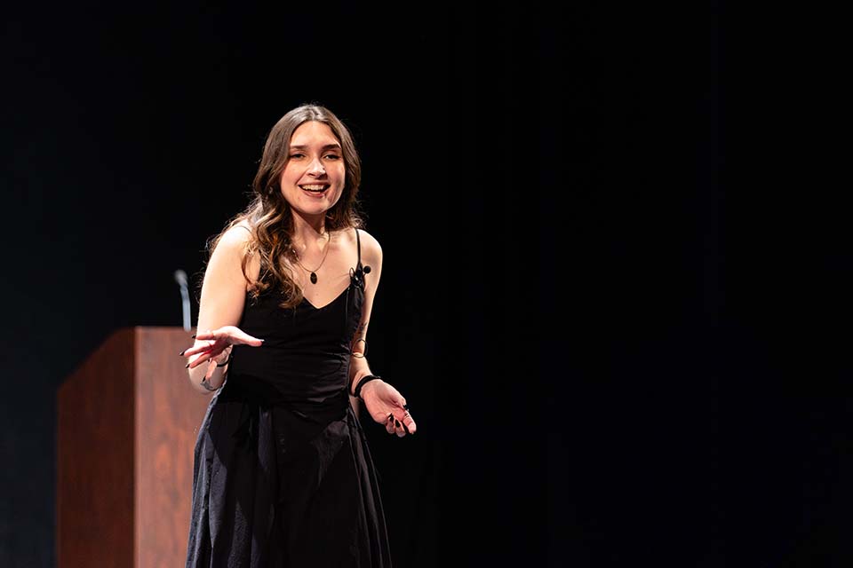 Dagny Michaels stands onstage presenting and holding out her hand.