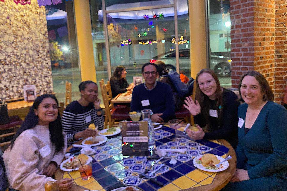 Students, alums, and staff sit together around a restaurant table.