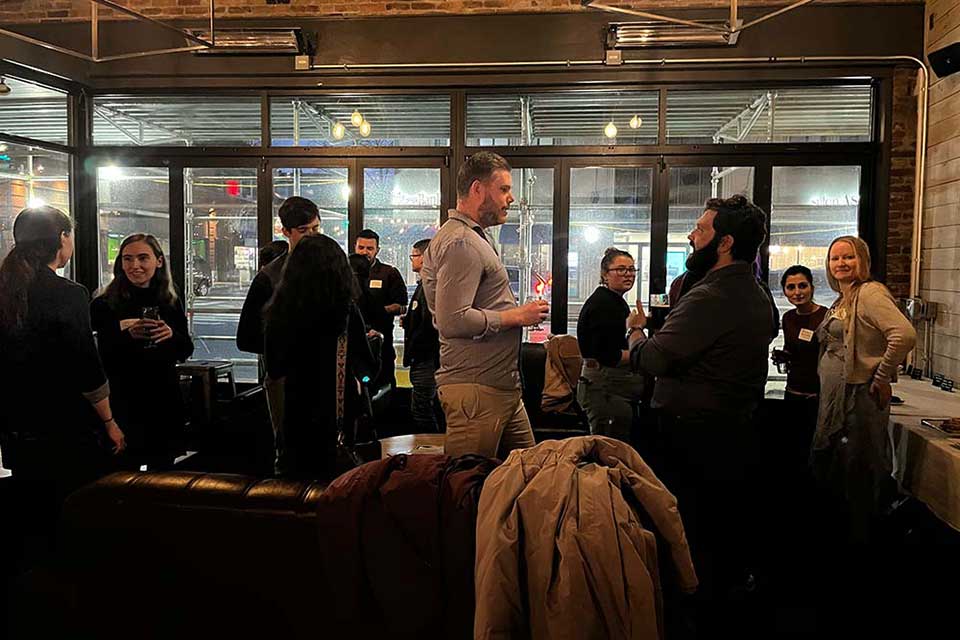 A group of people stand around chairs and tables in a restaurant, talking in small groups.