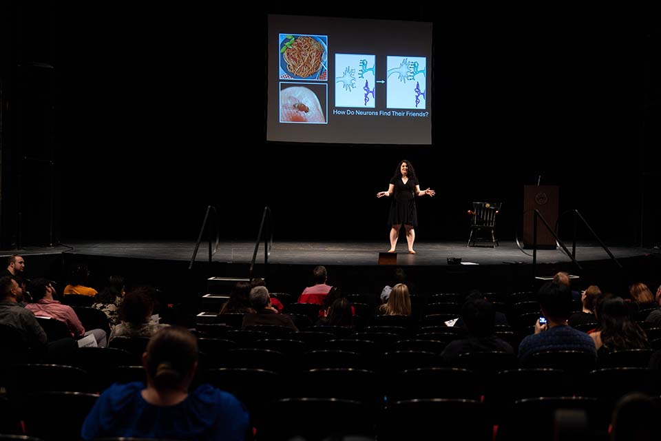 Catherine Carmona stands onstage, presenting in front of a slide, as the audience watches.