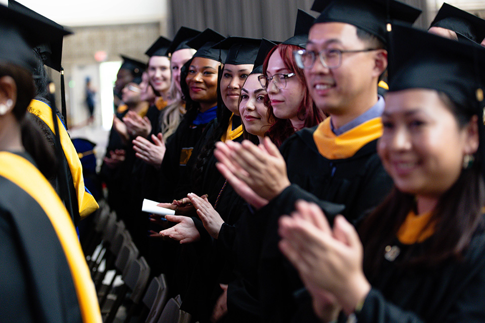 Students in graduation caps and gowns stand applauding.