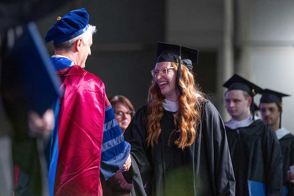 Dean Charles Golden shakes hands with a student; both wear regalia, and additional students in regalia are lined up to shake hands as well.
