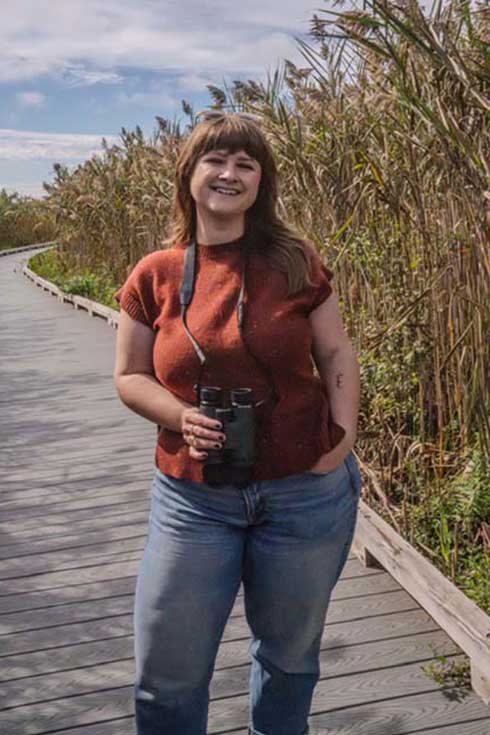 Jessi Brewer stands on a wooden walkway holding binoculars, next to some plants.