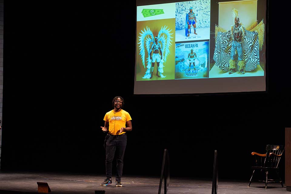 Kwame Gayle stands onstage, presenting in front of a slide showing men in colorful Carnival outfits.