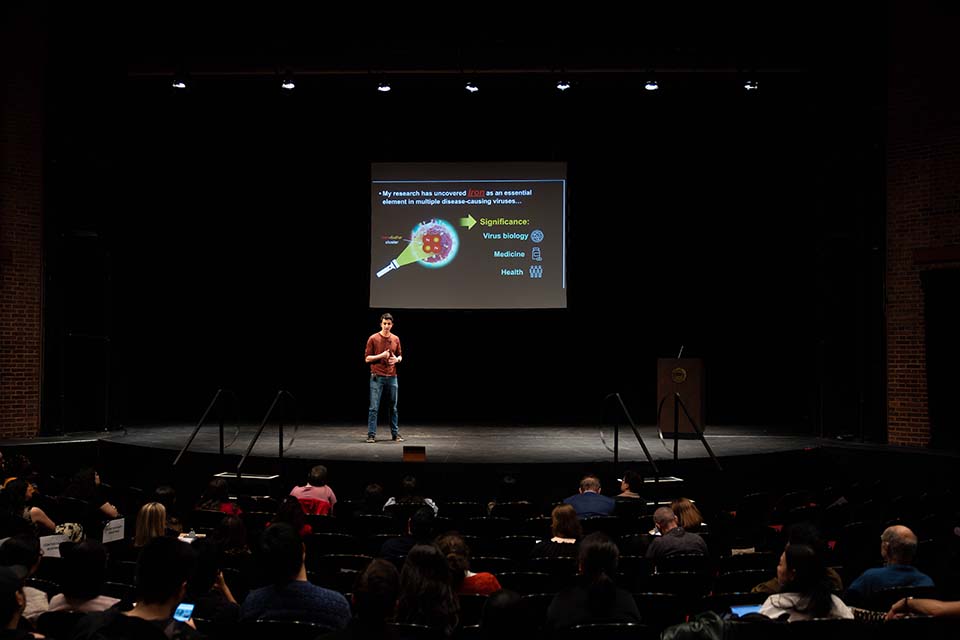 Trent Quist stands onstage, presenting in front of a slide, while the audience watches.