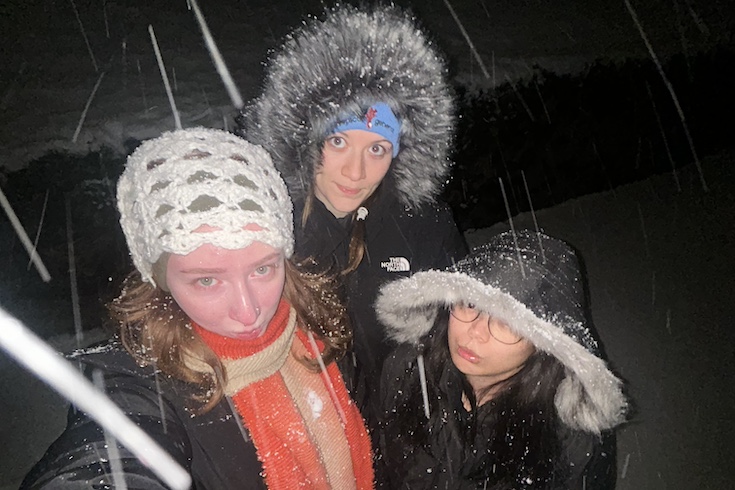 Mimi Kay, Mariko Tsuji, and Audrey Jordan, all heavily bundled up, look up at the camera as snow falls around them.