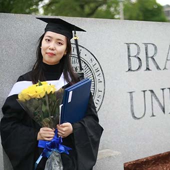 Wen Zhang stands in graduation robes by the Brandeis University sign.