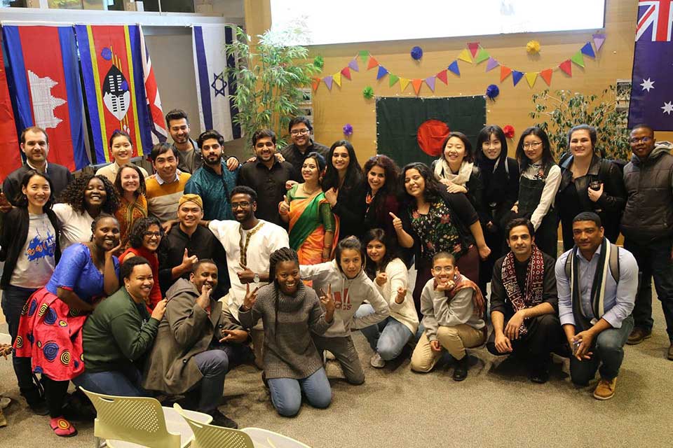 A large group of students stand in rows in front of various national flags.