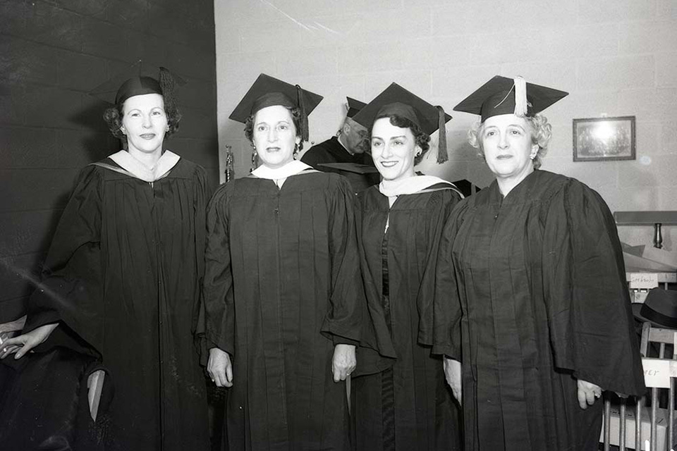 Four women in caps and gowns stand in a row.