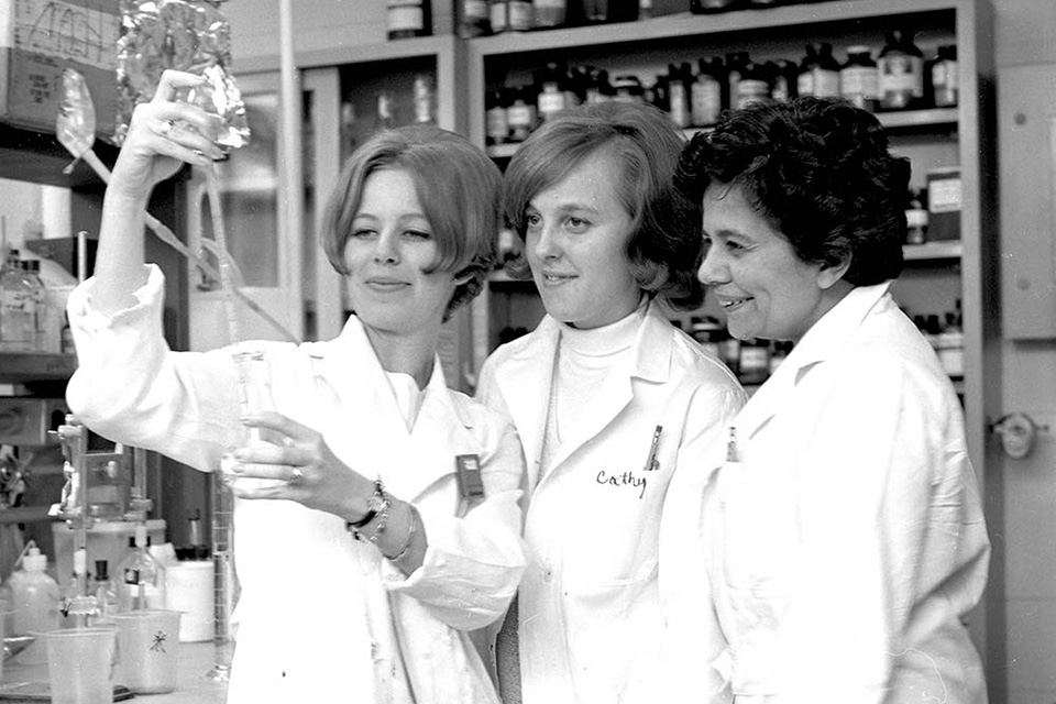 Three women in lab coats work in a lab with a test tube.