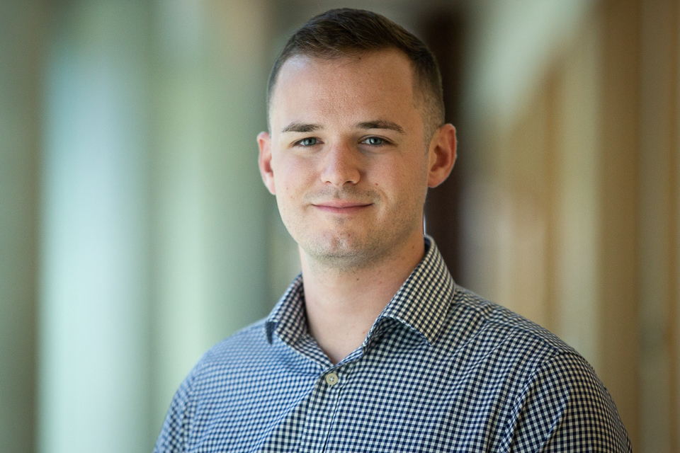 Headshot of Alex standing in front of a beige hallway