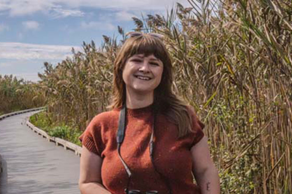 Jessi Brewer stands on a wooden walkway holding binoculars, next to some plants.