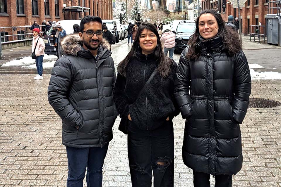Sayan Biswas, Jazmin Morales, and Marissa Ashton, all wearing winter coats, stand in a line in Brooklyn's DUMBO neighborhood.