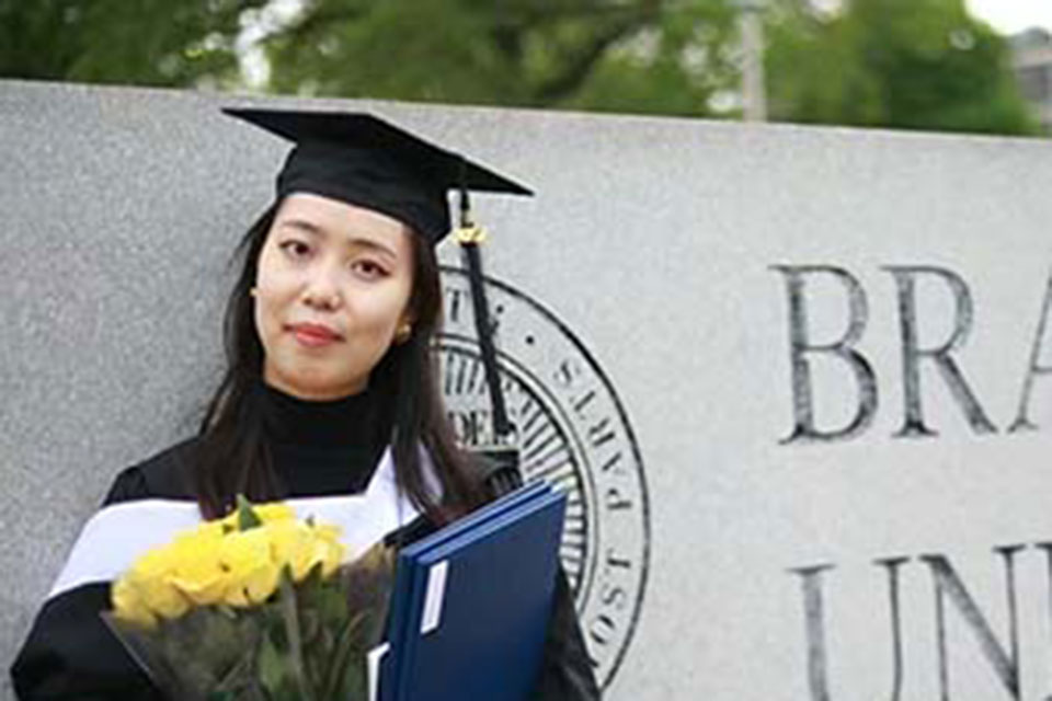 Wen Zhang stands in graduation robes by the Brandeis University sign.