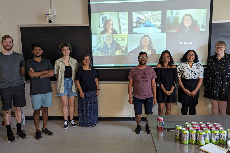 Eight members of the Women in Physics group stand in front of a blackboard, with six additional Zoom squares on a screen between them. There is a table with sodas in the foreground. 