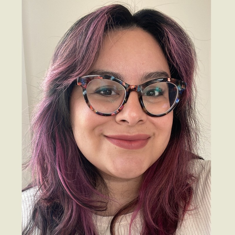 Alma smiling at the camera. She has faded magenta-streaked hair and large tortoise-shell glasses.