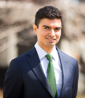 A headshot of Aneil, wearing a suit with a green tie and smiling at the camera.