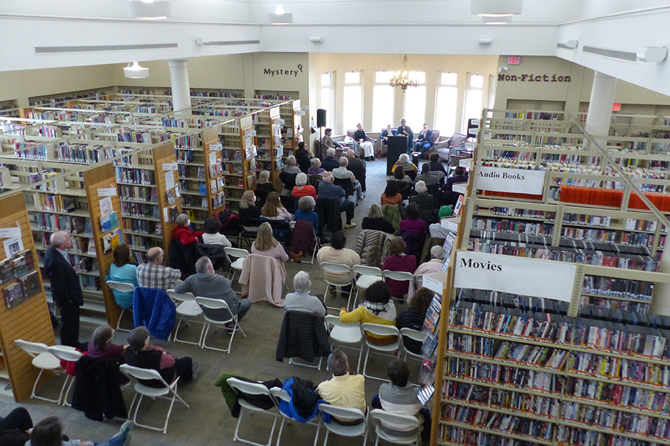 A group of people sit in chairs in a library facing presenters on a stage.